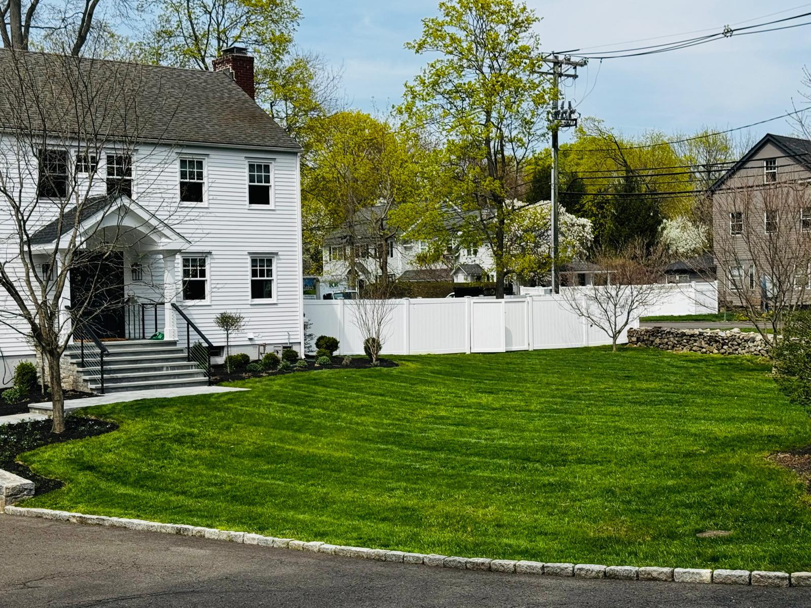 Striped lawn leading to a line of mature pines and a dark fence